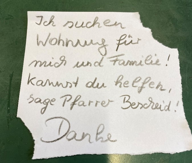 Ein Zettel, auf dem steht: 'Ich suche ein Wohnung für mich und Familie! Kannst du helfen, sage Pfarrer Bescheid! Danke.'